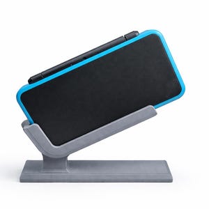 Könnte beinhalten: Eine schwarz-blaue Handheld-Spielkonsole wird auf einem grauen Ständer präsentiert. Die Konsole hat einen schwarzen Bildschirm und einen blauen Rahmen. Der Ständer ist grau und hat eine rechteckige Basis.