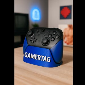 Könnte beinhalten: Ein schwarzer und blauer Nintendo Switch Pro Controller mit dem Wort GAMERTAG in Weiß. Der Controller steht auf einer Holzoberfläche. Der Hintergrund umfasst ein blaues Neonlicht und Kunstwerke.