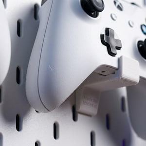 Könnte beinhalten: Weißer Xbox-Controller, montiert an einer weißen Lochwand mit einem 3D-gedruckten Halter. Der Controller hat schwarze Knöpfe und ein schwarzes Steuerkreuz. Der Halter trägt den Text "Made by 3000K.in".
