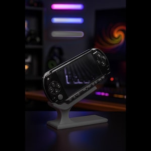 May include: A black Sony PSP handheld game console is displayed on a gray stand. The console has a black screen and buttons. The stand is a light gray color and has a modern design. The background is dark with neon lights.