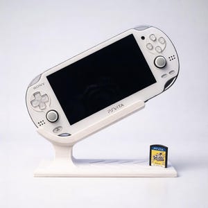 May include: A white Sony PS Vita handheld gaming console on a white stand, with a black screen and control buttons. A game cartridge labeled "PS Vita" and "PAC-MAN" is next to the console. The stand has the text "PSVITA" on it.