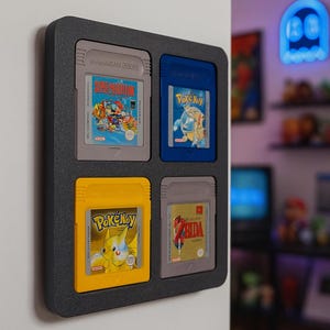 Könnte beinhalten: Eine schwarze Game Boy-Kassetten-Vitrine mit vier klassischen Game Boy-Spielen. Die Kassetten sind blau, gelb und grau und tragen Titel wie Super Mario Land und Pokémon.