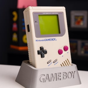 Könnte beinhalten: Ein cremefarbenes Nintendo Game Boy Handheld-Konsolen in einem grauen Ständer. Die Konsole hat einen grünen Bildschirm, ein schwarzes Steuerkreuz, rosa Knöpfe und den Text "Nintendo GAME BOY". Der Ständer ist mit "GAME BOY" graviert.