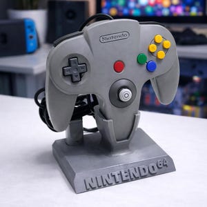 Könnte beinhalten: Ein grauer Nintendo 64 Controller auf einem passenden Ständer. Der Controller hat ein Steuerkreuz, eine rote A-Taste und farbige Knöpfe. Der Ständer ist mit "NINTENDO 64" beschriftet.