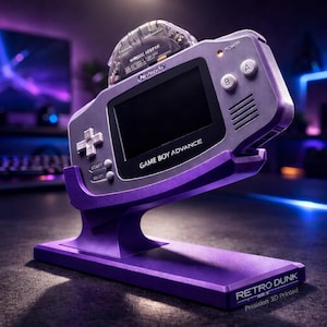 Può includere: Una console portatile Game Boy Advance viola esposta su un supporto viola abbinato. La console ha uno schermo nero e le parole "GAME BOY ADVANCE" sono stampate sulla parte anteriore. Una cartuccia di gioco è inserita nella parte superiore. Il supporto ha il testo "RETRO DUNK" e "Precision 3D Printed".