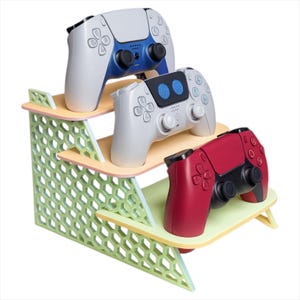 Può includere: Un supporto per controller di gioco a tre livelli con un design a nido d'ape verde. Il supporto contiene tre controller: bianco con dettagli blu, bianco con dettagli blu e rosso. Il supporto è in legno e dipinto con colori pastello.