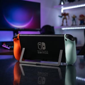 Pode incluir: Uma consola Nintendo Switch preta com comandos Joy-Con laranja e verde-azulado é exibida numa superfície refletora. A consola está num suporte prateado. O logótipo Nintendo Switch é visível.