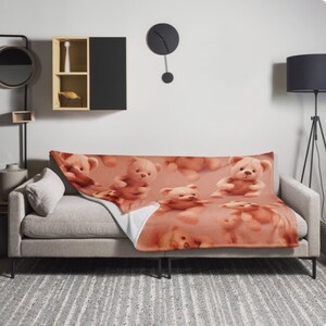 May include: A soft, plush throw blanket featuring a repeating pattern of brown teddy bears on a peach background. The blanket is draped over a light brown sofa.