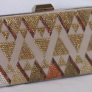 May include: Rectangular clutch with a geometric pattern of gold, bronze, and silver rhinestones. The clutch has a silver metal frame and clasp. The pattern consists of triangles and diagonal lines, creating a sparkling, eye-catching accessory.