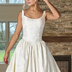 May include: A white wedding dress with a floral pattern. The dress features a fitted bodice with a square neckline and wide straps. The skirt is full and flowing. The background includes a window and a brick fireplace.