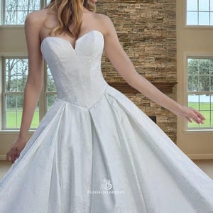 May include: A white strapless wedding dress with a sweetheart neckline and a full skirt. The bodice has a textured floral pattern, and the skirt is pleated. The dress is suitable for a formal event.