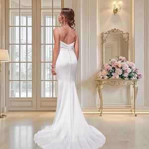 Op de afbeelding: Een witte, vloerlange bruidsjurk met een getailleerd silhouet en een lichte zeemeermin-flare. De jurk heeft spaghettibandjes, een lage rug en een rij knopen op de rug. De tekst "BLISSFULLBRIDES" is zichtbaar onderaan de jurk.
