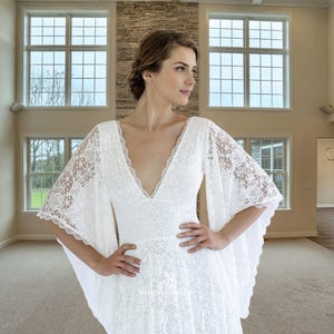 May include: A white lace wedding dress with a plunging V-neckline and wide, flowing sleeves. The dress features a floral lace pattern and a fitted bodice. The model is standing in a large room with windows.