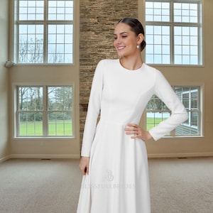 Könnte beinhalten: Ein weißes, langärmliges Kleid mit rundem Ausschnitt und ausgestelltem Rock. Das Kleid hat ein figurbetontes Oberteil und eine dezente Nahtdetail. Das Logo "Blissfull Brides" ist auf dem unteren Teil des Kleides sichtbar.