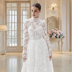 May include: A white lace wedding dress with long sleeves and a high neckline. The dress features intricate floral lace detailing and a fitted bodice. The skirt flows to the floor. The dress is worn in a well-lit room with a mirror and flowers in the background.