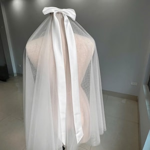 Puede incluir: Un velo de novia blanco con un lazo y cintas de satén. El velo está hecho de una tela transparente y ligera que cae sobre los hombros. El lazo está colocado en la parte superior del velo, añadiendo un toque de elegancia.