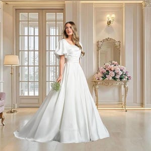 May include: A white satin wedding dress with a square neckline and short, puffed sleeves. The dress features a fitted bodice and a flowing A-line skirt. The bride is holding a small bouquet of greenery. The background includes a gold mirror and a table with flowers.