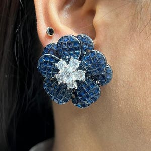 Op de afbeelding: Een close-up van een bloemvormige oorbel. De oorbel heeft een donkerblauw, getextureerd bloemblaadjesontwerp met een cluster van heldere stenen in het midden. De oorbel wordt gedragen op het oor van een persoon.