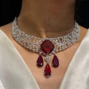 Peut inclure: Un collier argenté orné d'un motif floral et d'une grande pierre rouge foncé carrée au centre. Trois gemmes rouges en forme de goutte pendent en dessous. Le texte "PREMIUM QUALITY" est visible.