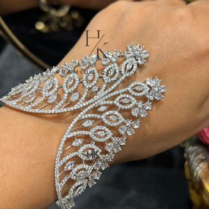 Puede incluir: Un brazalete plateado con un diseño abierto inspirado en hojas, incrustado con numerosas piedras transparentes de corte marquesa y redondo. La pieza muestra un patrón simétrico ramificado con detalles florales en los extremos y el texto HK PREMIUM QUALITY.