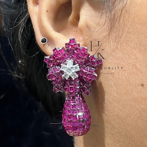 Puede incluir: Primer plano de un pendiente magenta con forma floral y colgante de lágrima. El pendiente está incrustado con pequeñas piedras preciosas de corte cuadrado. Una pequeña gema blanca en forma de estrella está en el centro del diseño floral.