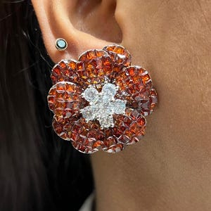 Op de afbeelding: Een grote, bloemvormige oorbel met een centrale cluster van witte edelstenen en bloemblaadjes bestaande uit kleine, vierkante, oranje-bruine stenen. Een kleinere, ronde oorbel met een blauwgroene steen is ook zichtbaar.