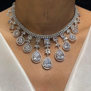 Puede incluir: Un collar choker plateado con una doble fila de piedras de circonita cúbica y una fila colgante de piedras de circonita cúbica en forma de pera. El collar tiene una etiqueta "H/K PREMIUM QUALITY".