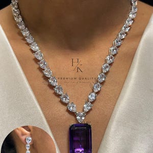 Pode incluir: Um conjunto de colar e brincos de prata. O colar apresenta um grande pendente retangular de pedra preciosa roxa escura. O colar e os brincos têm pedras claras em forma de pêra. Os brincos espelham o design do colar, com uma pedra roxa a condizer.