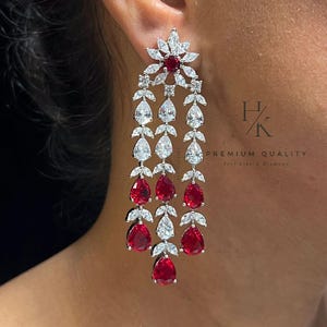 Peut inclure: Boucles d'oreilles pendantes avec un motif floral au sommet, avec une pierre rouge centrale entourée de pierres blanches. Les boucles d'oreilles sont composées de pierres rouges et blanches en forme de goutte. Le texte "PREMIUM QUALITY" est visible.