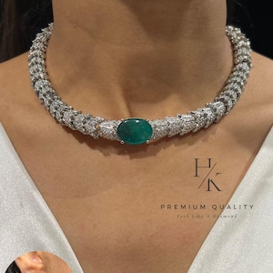 May include: A silver-toned necklace with a large oval green gemstone at the center. The necklace is encrusted with small, sparkling stones. A matching earring is visible in the lower left corner. The text "PREMIUM QUALITY" is also visible.