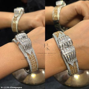 May include: A hinged bracelet featuring a central row of baguette-cut clear stones flanked by triangular sections of champagne-colored stones, all set in a silver-toned metal frame with pavé-set clear stones along the edges and a logo mark.