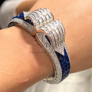 Puede incluir: Una pulsera plateada con un diseño geométrico. La pulsera presenta una combinación de piedras preciosas claras y azules. Las piedras claras son rectangulares y las azules son cuadradas. La pulsera se lleva en la muñeca.