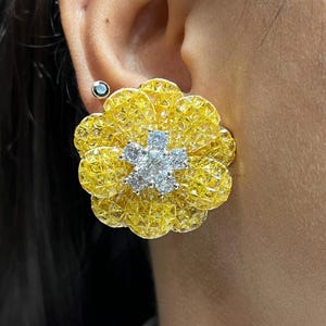Op de afbeelding: Close-up van een gele bloemvormige oorbel met een cluster van witte edelstenen in het midden. De oorbel heeft een getextureerd oppervlak en wordt op het oor gedragen. Een kleine, ronde oorbel is ook zichtbaar.
