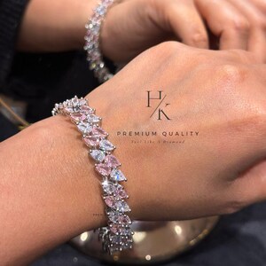 Pode incluir: Uma pulseira prateada adornada com pedras em forma de pêra rosa e transparentes alternadas. A pulseira está em um pulso, com outra pulseira visível ao fundo. O texto "HK PREMIUM QUALITY Feel Like A Diamond" também é visível.
