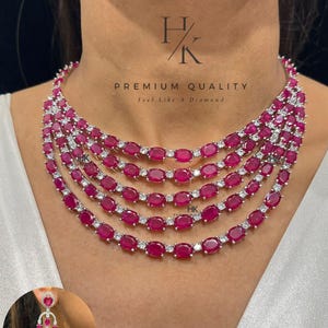 Può includere: Una collana multistrato con pietre color rubino taglio ovale e piccole pietre bianche. La collana è a strati e indossata intorno al collo. È visibile anche un paio di orecchini abbinati con pietre color rubino.