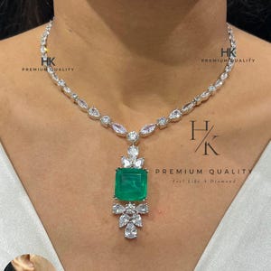 Pode incluir: Um colar de prata com um grande pingente quadrado de pedra preciosa verde esmeralda, rodeado por pedras preciosas claras menores. O colar tem uma corrente delicada com pedras preciosas claras alternadas. Um brinco a condizer também é visível. O texto na imagem diz "HK PREMIUM QUALITY" e "Feel Like A Diamond".