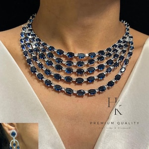 Può includere: Un set di collana e orecchini abbinati. La collana presenta file di pietre preziose ovali blu scuro intervallate da pietre più piccole e trasparenti. Gli orecchini hanno un design simile, con una singola pietra blu.