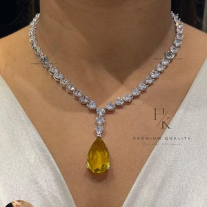 May include: A stunning necklace with a yellow pear-shaped pendant. The necklace is adorned with clear, heart-shaped stones set in a silver-toned metal. Matching earrings are visible. The text "PREMIUM QUALITY" is visible. The necklace is a beautiful piece of jewellery.