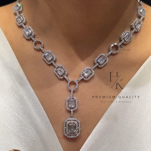 Peut inclure: Un collier argenté orné de pierres rectangulaires taille émeraude entourées de petites pierres pavées. Le design comprend un pendentif central avec deux maillons rectangulaires superposés. Le texte HK Premium Quality Feel Like A Diamond est visible.