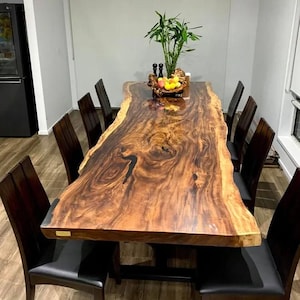 May include: A large, live-edge wooden dining table with a rich, dark brown finish. The table is surrounded by ten dark wooden chairs with black leather seats. A decorative centerpiece of fruit and greenery sits on the table.