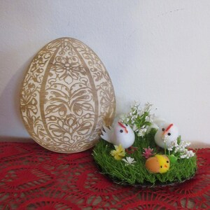 May include: A large wooden Easter egg with a detailed floral pattern is displayed beside a small decorative nest. The nest contains two white chicks, flowers, and a small yellow and orange chick. The scene is set on a red lace tablecloth.
