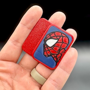 May include: A rectangular red plastic pin featuring a raised profile graphic of a superhero mask in red and black with white eye lenses. The pin has a textured red background section and a smooth blue section surrounding the character illustration.