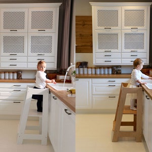 May include: Two children stand on wooden step stools in a white kitchen. The children are wearing casual clothing and are reaching for the sink. The kitchen has white cabinets and a wooden countertop.