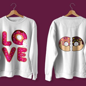 K&ouml;nnte beinhalten: Zwei wei&szlig;e Sweatshirts mit Donut-Designs. Ein Sweatshirt hat das Wort "LOVE" mit Donut-Buchstaben ausgeschrieben. Das andere Sweatshirt hat zwei halb gegessene Donuts, einen rosa und einen Schokoladen-Donut.