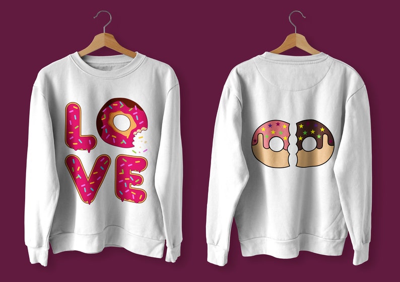 K&ouml;nnte beinhalten: Zwei wei&szlig;e Sweatshirts mit Donut-Designs. Ein Sweatshirt hat das Wort "LOVE" mit Donut-Buchstaben ausgeschrieben. Das andere Sweatshirt hat zwei halb gegessene Donuts, einen rosa und einen Schokoladen-Donut.