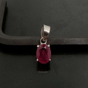 Peut inclure: Un pendentif en argent avec une grosse pierre précieuse rouge foncé de forme ovale. La pierre précieuse est sertie dans une lunette en argent et a une surface polie. Le pendentif a un design simple et élégant. Le texte "PKJEWELERS" est en bas.