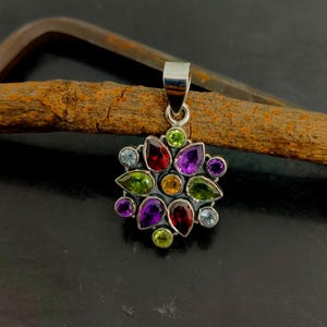 以下が含まれることがあります： 花柄に配置されたマルチカラーの宝石のクラスターを特徴とするシルバーペンダント。デザインには、ペアシェイプおよびラウンドカットのガーネット、アメジスト、ペリドット、シトリン、ブルートパーズが含まれており、暗い金属製のフレームにセットされています。