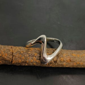 Op de afbeelding: Een gepolijste zilveren ring met een uniek, abstract ontwerp. De ring heeft een golvende zigzagvorm die omhoog buigt. De ring wordt getoond op een bruine houten stok tegen een donkere achtergrond.