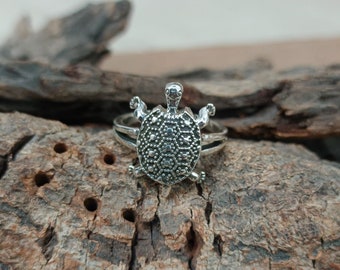 May include: A silver turtle ring with a detailed design. The ring is on a piece of weathered wood.