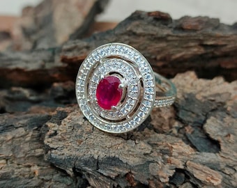 May include: A silver ring featuring a central oval-cut red gemstone surrounded by two concentric halos of small clear stones. The metal band is also encrusted with small clear stones. The text pkjeweler is visible at the bottom of the image.